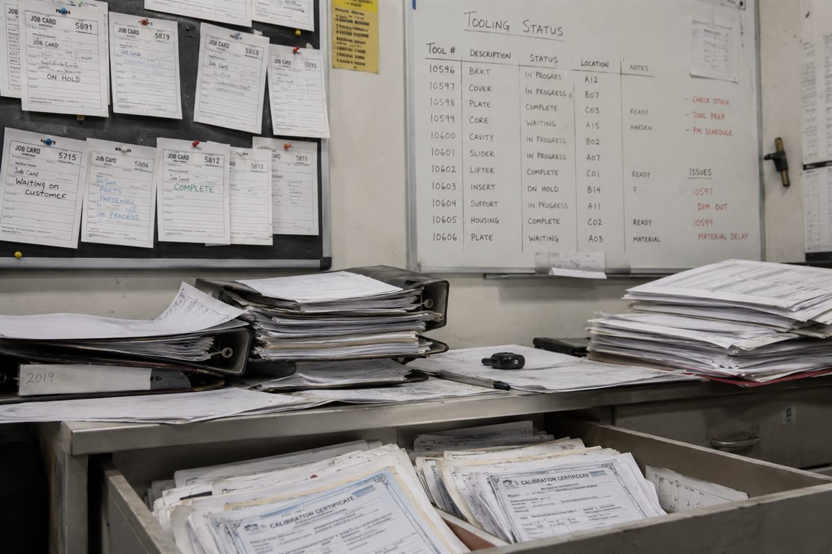 A toolroom desk buried in paper job cards, a handwritten tooling-status whiteboard, binders of records, and a drawer of loose calibration certificates.
